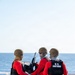 Three U.S. Coast Guard personnel are stationed on the deck of a vessel at sea, gathered around a mounted weapon system. They are wearing red long-sleeve shirts, navy blue pants, tactical vests, ballistic helmets, and face coverings. The scene appears to depict a weapons training or readiness drill conducted on open water.