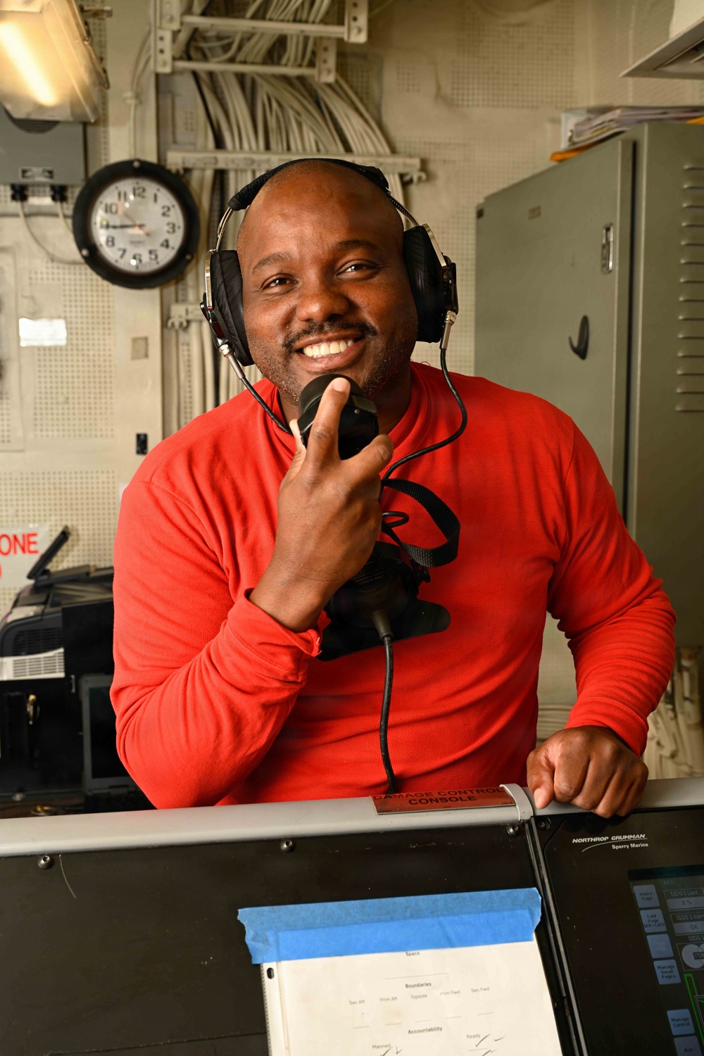 A smiling man wearing a red long-sleeve shirt and headset is seated at a Damage Control Console aboard what appears to be a naval vessel. He is holding a microphone and appears to be in the middle of a communication task. The setting is an interior shipboard operations room with visible equipment, cables, and a wall clock.