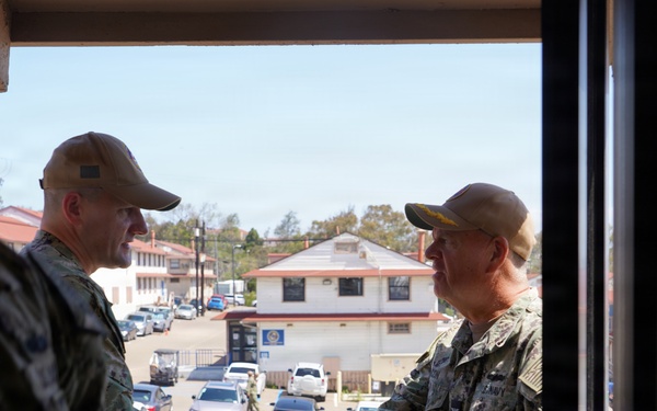 Two military personnel in U.S. Navy/military camouflage uniforms are shaking hands on what appears to be an elevated balcony or landing of a military installation building. The scene is photographed from inside a doorway looking outward, with a military base compound visible in the background including vehicles, buildings, and a parking area. The setting appears to be a formal or semi-formal greeting between service members.
