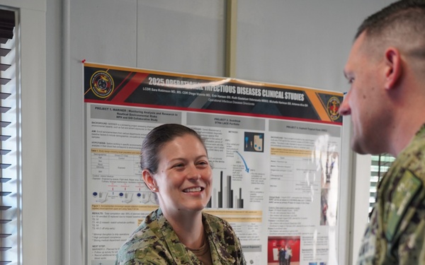 Two U.S. Navy personnel in NWU Type III (green camouflage) uniforms are shaking hands in an indoor office setting. The female service member, identified by her name tape as 'Robinson,' is smiling broadly. Behind her is a research poster titled '2025 Operational Infectious Diseases Clinical Studies.'