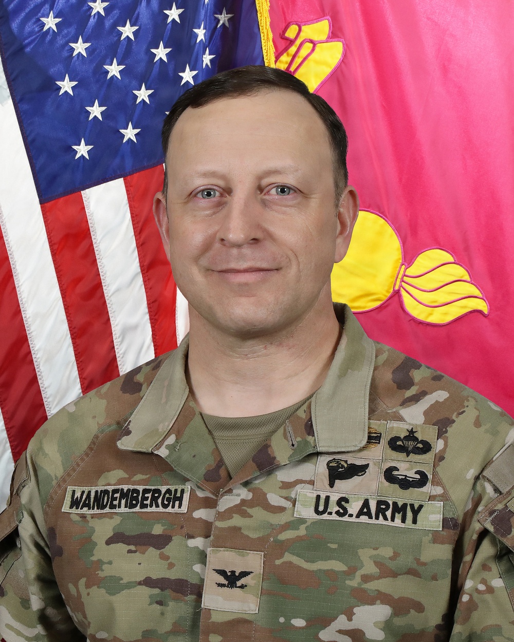 A U.S. Army Colonel in an official military portrait, wearing Operational Camouflage Pattern (OCP) uniform with the name tape 'WANDEMBERGH' and 'U.S. ARMY' tape visible. The subject is posed in front of an American flag and what appears to be a unit or branch guidon/flag with yellow and pink coloring. Multiple qualification badges are visible on the uniform.