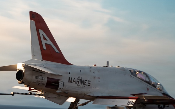 A T-45 Goshawk trainer jet marked with 'MARINES' and bureau number 165623 is landing on an aircraft carrier deck, with the arresting hook engaged on the wire. The aircraft belongs to Training Wing One (TW-1), VT-9 squadron, and bears the modex number 181 with a distinctive red-and-white color scheme. The photo is taken at dusk or dawn, with warm ambient lighting illuminating the scene.