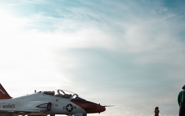 A U.S. Marines T-45 Goshawk trainer jet (bureau number 160) is positioned on the flight deck of an aircraft carrier. The aircraft features a distinctive red and white color scheme with a large star-and-bar insignia and 'MARINES' lettering on the fuselage. Several flight deck crew members in green jerseys are visible observing the aircraft, suggesting pre-launch or post-landing operations.