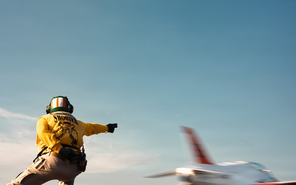 A naval flight deck crew member, identified as a 'Shooter' by the text on their yellow jersey, signals the launch of a jet aircraft from an aircraft carrier deck. The aircraft, appearing to be a T-45 Goshawk or similar trainer/fighter jet in white and red livery, is captured mid-launch with motion blur indicating high speed. The low-angle perspective dramatically emphasizes both the crew member's dynamic launch posture and the aircraft's acceleration.
