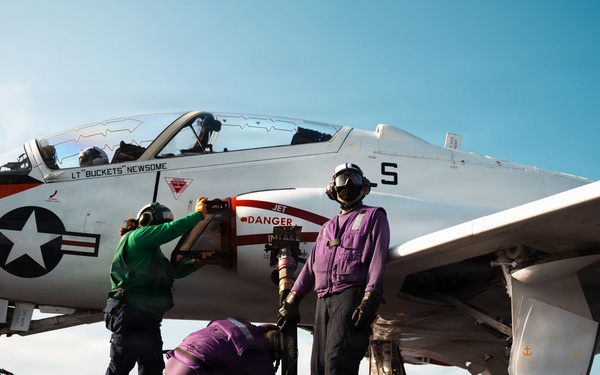U.S. Navy deck crew personnel are conducting pre-flight or maintenance operations on a carrier-based jet aircraft on what appears to be an aircraft carrier flight deck. The aircraft displays U.S. Navy markings including a roundel, orange accent colors, and the name 'LT BUCKETS NEWSOME' on the canopy rail. Three crew members in colored jerseys are working around the aircraft's nose and landing gear area.