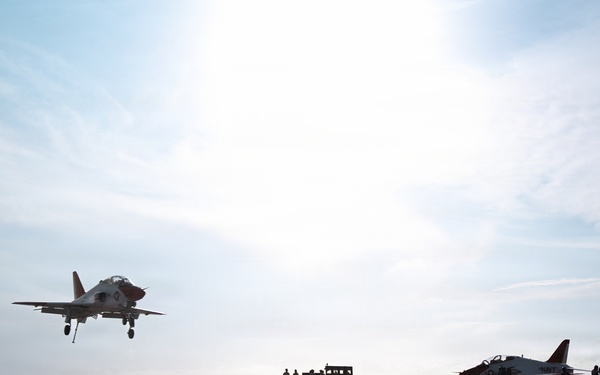 A military jet aircraft is captured on approach or just landing on what appears to be an aircraft carrier flight deck. Two additional aircraft are parked on the deck to the right, with several personnel visible in silhouette. The image is taken from a low angle against a bright, hazy sky, creating strong silhouette contrast.