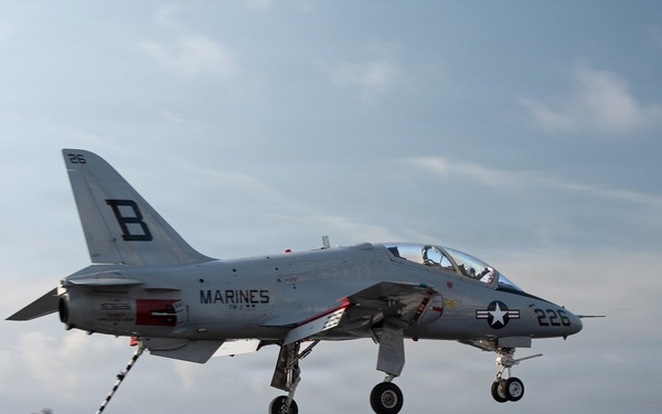 A U.S. Marines aircraft, marked with the number 226 and letter 'B', is captured in the process of landing or taking off from an aircraft carrier deck. The jet, identifiable as a McDonnell Douglas AV-8B Harrier II based on its distinctive profile, has its landing gear fully deployed. The image was taken from the carrier deck level, with open ocean visible in the background under a partly cloudy sky.