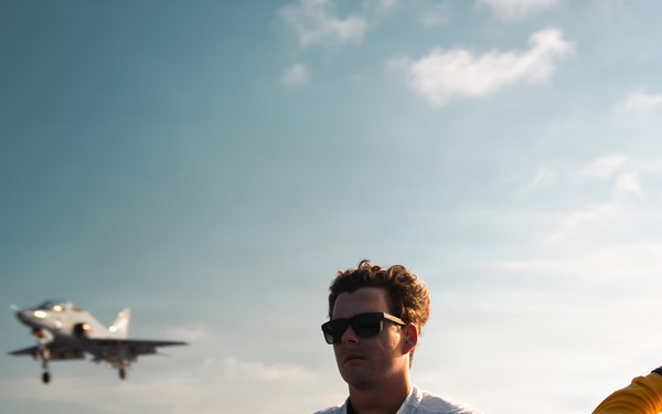 Two personnel are standing on the flight deck of an aircraft carrier, with a military jet aircraft visible in the background either launching or landing. The individual in the foreground wears a white vest with the number '339' and a US flag patch, while the person to the right wears a yellow flight deck jersey and appears to be communicating via a headset. The scene is set over open ocean under a partly cloudy sky.