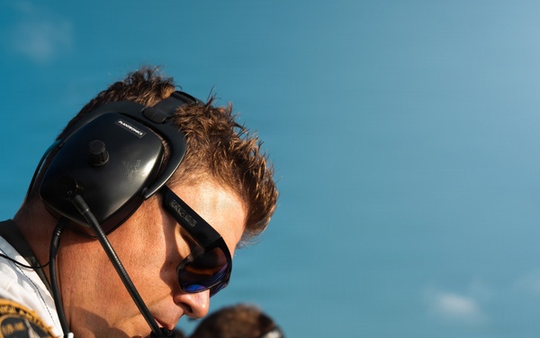 A close-up photograph of a male aviation or military personnel wearing a flight suit, communication headset, and reflective sunglasses. He appears to be focused on a task, leaning over a surface, with at least one other similarly dressed individual visible in the background. The image is taken from a low angle against a clear blue sky.
