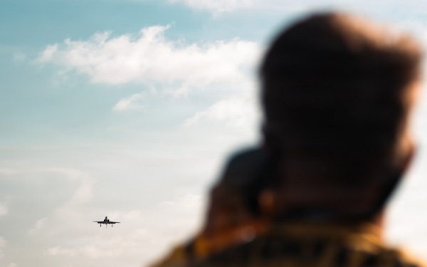 A person wearing a yellow flight deck jersey stands on what appears to be an aircraft carrier deck, watching a military jet aircraft in the distance over open ocean. The aircraft is silhouetted against a partly cloudy sky, appearing to be either launching or departing from the carrier. The foreground figure is intentionally blurred using shallow depth of field, drawing focus to the aircraft on the horizon.