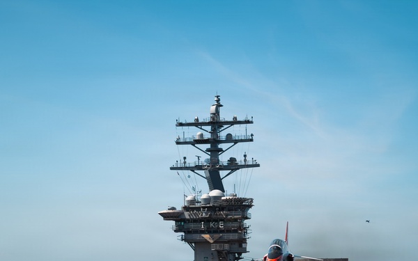 The image shows the flight deck of a U.S. Navy aircraft carrier, identifiable by the letters 'IKE' on the island superstructure, indicating USS Dwight D. Eisenhower (CVN-69). Multiple red/orange training jets, likely T-45 Goshawks, are visible on deck with flight deck crew in colored jerseys performing operations. The scene captures active flight deck operations under clear blue skies at sea.
