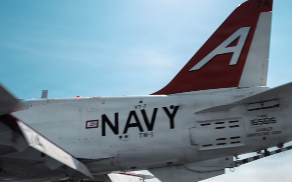 A U.S. Navy T-45 Goshawk trainer jet is captured in motion on what appears to be an aircraft carrier flight deck, likely during a landing or takeoff operation. The aircraft displays distinctive red and white tail markings with a large letter 'A' and bears the bureau number 165616. The low-angle, dynamic shot conveys speed and motion blur consistent with carrier deck operations.