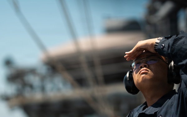 A naval service member in dark blue utility uniform stands on the deck of a large warship, shielding their eyes from the sun while looking upward. The sailor is equipped with hearing protection (ear defenders) and holds a small object, likely a radio or tool. The background reveals the superstructure of what appears to be a large naval vessel, possibly an aircraft carrier, with open ocean visible.