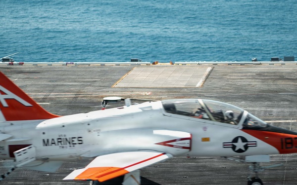 A U.S. Marines T-45 Goshawk trainer jet (bureau number 183) is captured during a carrier landing or catapult launch on an aircraft carrier flight deck. The aircraft features the distinctive orange-and-white training color scheme with a large 'A' on the tail fin. Smoke is visible near the main landing gear, consistent with either a arrested landing tire contact or catapult launch activity.
