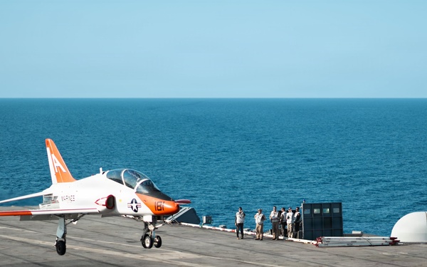 A U.S. Navy T-45 Goshawk trainer jet in distinctive orange-and-white training livery is landing or taking off on an aircraft carrier flight deck at sea. The aircraft bears the number '161' and MARINES markings, with a second aircraft marked '106' visible on the right side of the deck. Several flight deck personnel are observed standing near the deck edge observing the operation.