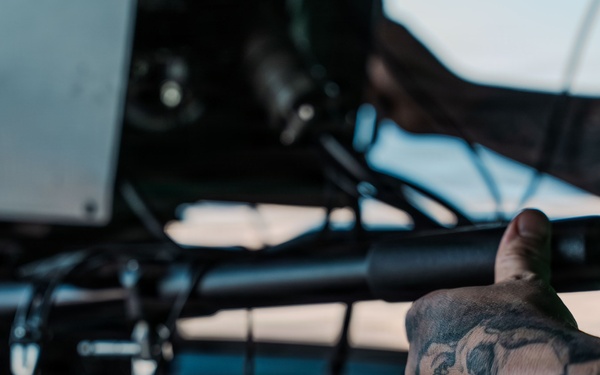 A close-up photograph showing a heavily tattooed forearm and hand gripping what appears to be a mounted weapon or mechanical equipment. The tattoos on the arm include a skull and rose motifs in black ink. The setting appears to be inside or near a vehicle or aircraft, with headphones and mechanical components visible in the background.