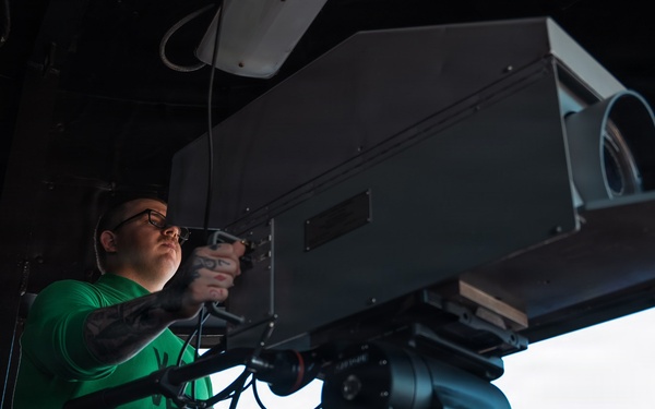 A tattooed individual wearing a green t-shirt and glasses is operating or adjusting a large professional broadcast camera mounted on a Cartoni tripod head. The scene appears to be inside a stadium press box or elevated broadcast position, with an open view to the outside. The low-angle shot emphasizes the scale of the professional camera equipment.