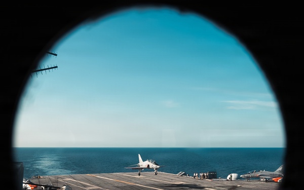 The image is captured through a circular porthole window aboard a naval aircraft carrier at sea. A jet aircraft, consistent with a Douglas A-4 Skyhawk design, is visible on the flight deck in what appears to be a takeoff or landing roll. Additional parked aircraft, deck crew personnel, and carrier equipment are visible on the deck under clear blue skies.