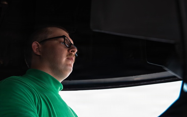 A young man wearing black-framed glasses and a green sweatshirt is seated inside what appears to be a large vehicle, likely a truck or heavy equipment cab. He is looking forward through the windshield, suggesting he may be driving or preparing to drive. The image is taken from a low angle inside the cab, creating a dramatic perspective.