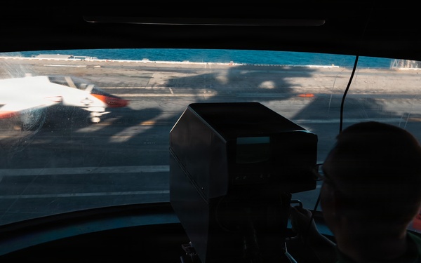 A person in a green shirt is visible in silhouette inside what appears to be a control tower or observation booth on an aircraft carrier flight deck. Through the curved panoramic window, a jet aircraft with orange and white markings is visible on the deck, possibly during launch or taxi operations. A second aircraft with US military markings is visible to the right side of the frame.