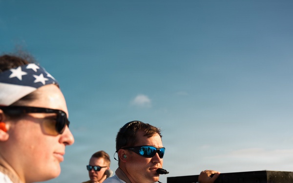 The image shows several military personnel, likely U.S. Navy sailors, on what appears to be the deck or bridge area of a naval vessel. They are wearing flight deck gear including tan/khaki uniforms, white float coats, and ear protection. One individual is speaking into a communication device while operating equipment.