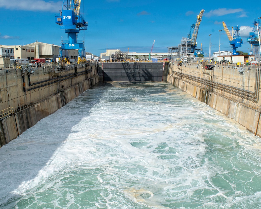 The image shows a large dry dock being flooded with turbulent water, likely in preparation for receiving or releasing a vessel. The facility appears to be a naval or commercial shipyard, identifiable by the large blue cranes labeled 'P-76' and yellow cranes visible in the background. The concrete dock walls and caisson gate at the far end are clearly visible as water rushes in with significant force.