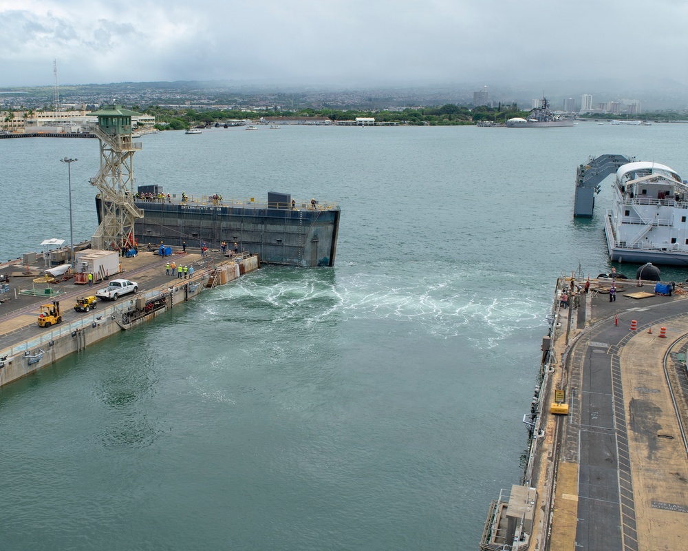 An aerial view of a naval shipyard facility, likely Pearl Harbor, Hawaii, showing a large floating dry dock being maneuvered or flooded at a pier. Workers in high-visibility vests are visible on the dock and surrounding quay areas, with turbulent water indicating active ballasting or de-ballasting operations. A gray naval vessel is moored to the right, and what appears to be the USS Missouri battleship memorial is visible in the background across the harbor.
