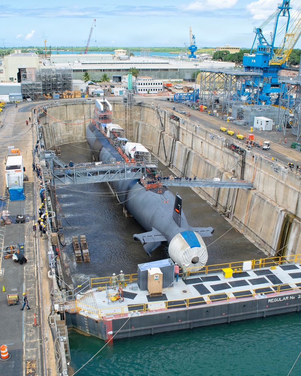A large military submarine is positioned in a dry dock facility, partially flooded, undergoing maintenance or inspection operations. The facility appears to be a naval shipyard with multiple cranes, scaffolding, and support structures visible. Personnel in safety gear are present on walkways and platforms surrounding the vessel.