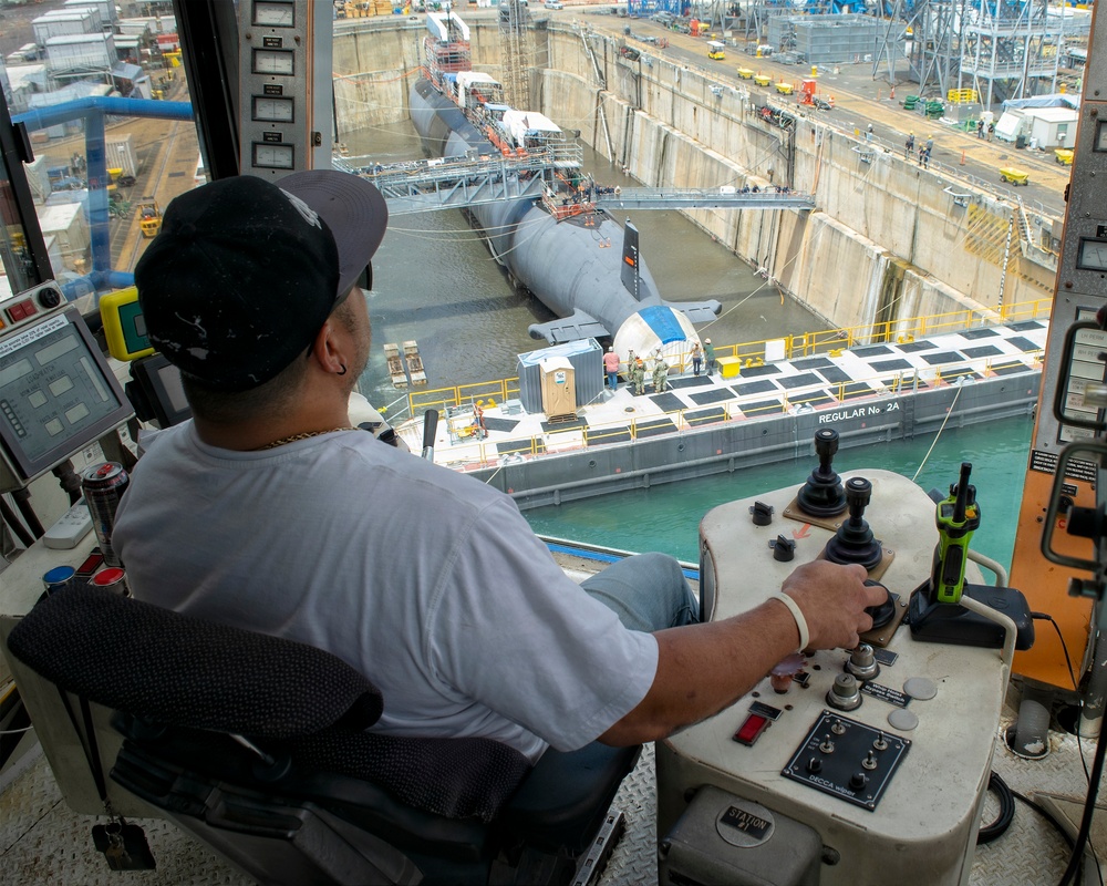 A crane or dock equipment operator is seated at a control console inside an elevated cab, overseeing a dry dock facility where at least two submarines are undergoing maintenance or refit. A barge labeled 'REGULAR No. 2A' is positioned alongside the submarines in the dry dock basin. The facility appears to be a busy naval shipyard with numerous workers, vehicles, and support structures visible.