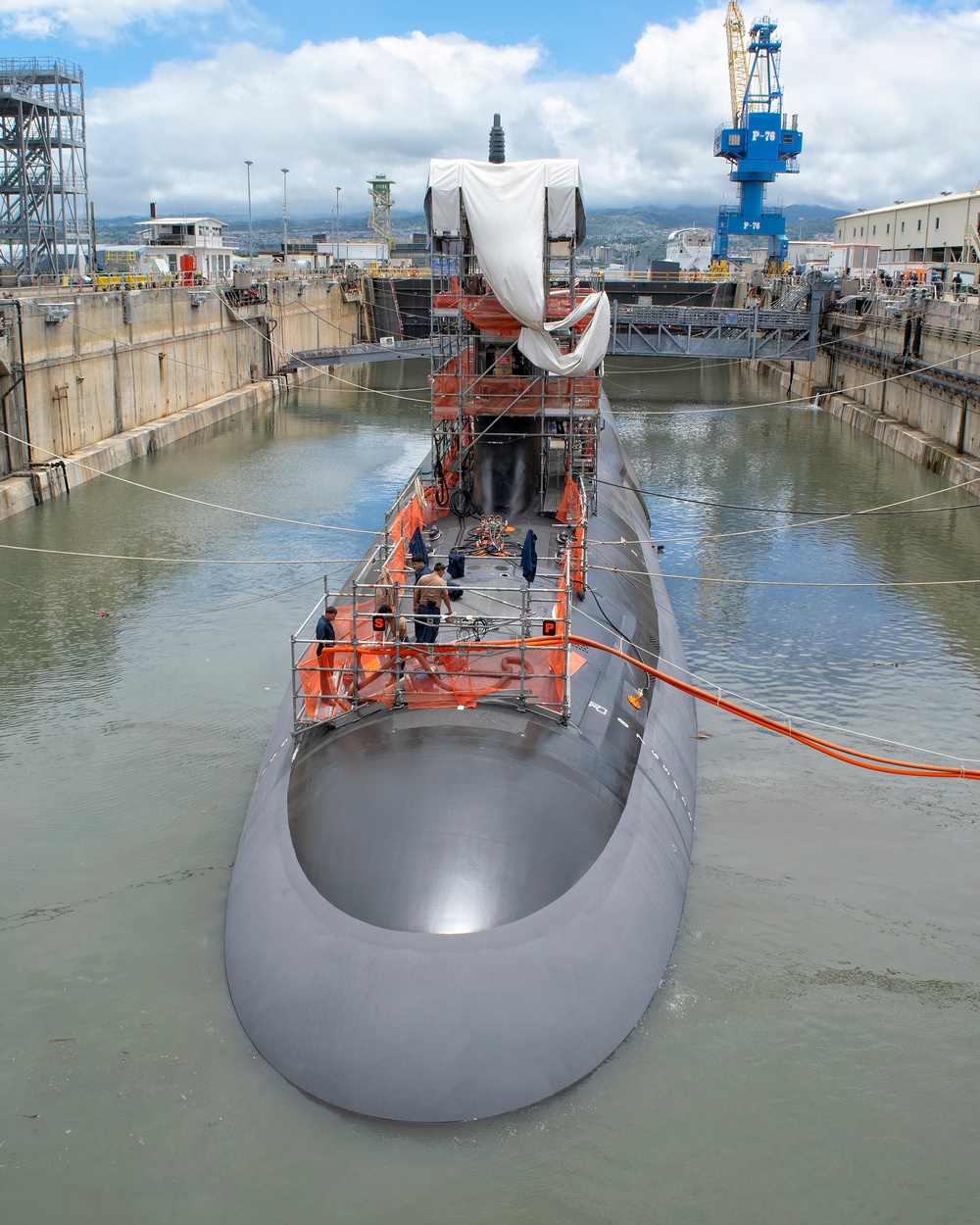 A military submarine is undergoing maintenance or refit in a dry dock facility, with the dock partially flooded. Workers in navy uniforms are visible on scaffolding platforms atop the submarine's hull, and the sail (conning tower) is partially covered with a protective tarp. The facility appears to be a naval shipyard, identifiable by the blue crane marked 'P-76' in the background.
