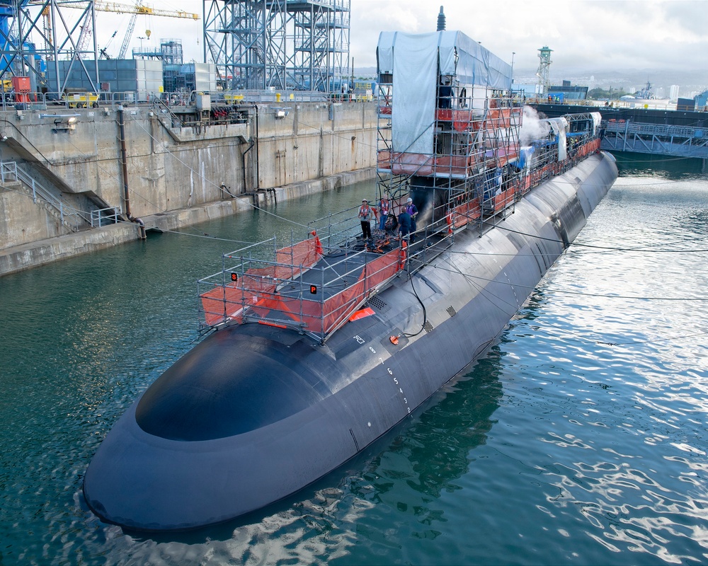 A large military submarine is moored alongside a dry dock or naval shipyard facility, undergoing maintenance or refit work. Scaffolding and temporary work platforms with orange safety barriers are erected on the vessel's hull and sail area, with a tarpaulin covering part of the upper structure. Several workers are visible on the deck, and steam or vapor is visible near the sail, suggesting active mechanical work.