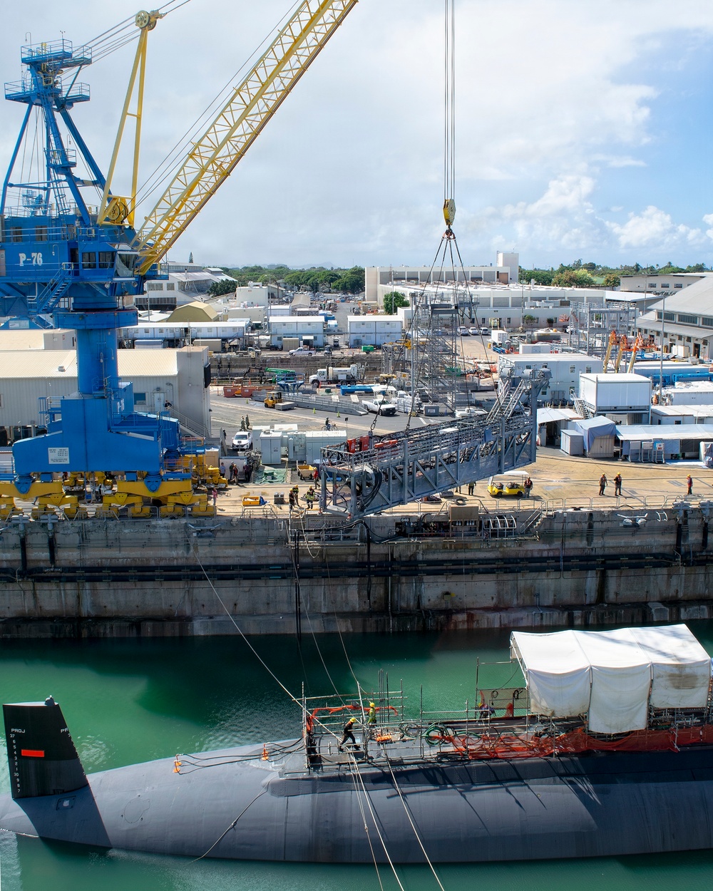 The image shows a naval shipyard facility with a submarine moored alongside a dry dock or pier undergoing maintenance work. A large blue gantry crane labeled 'P-76' with a yellow boom is actively lifting a large steel scaffolding or work platform structure. The facility appears to be a U.S. Navy installation, likely Pearl Harbor Naval Shipyard in Hawaii, based on the tropical vegetation and architectural style visible in the background.