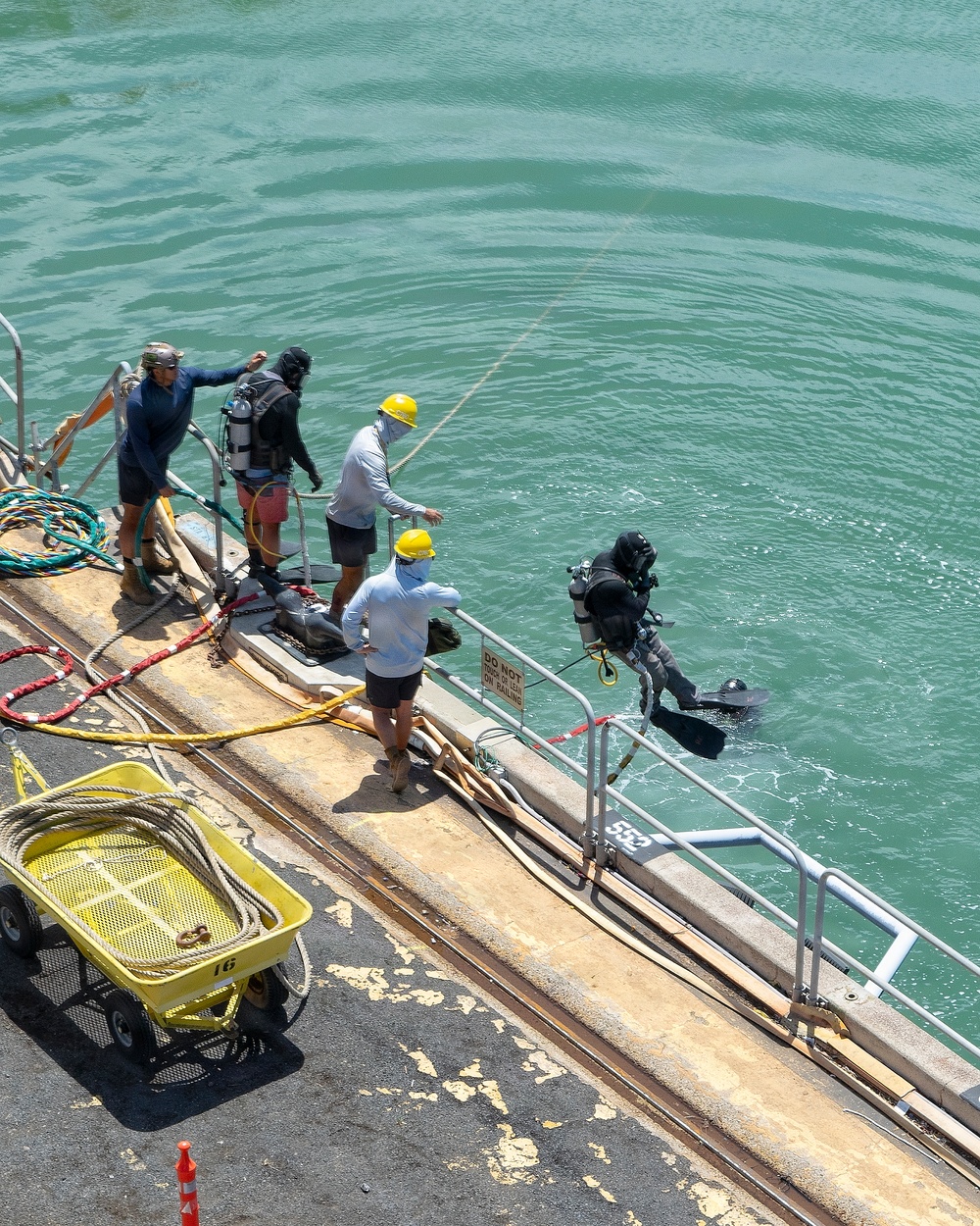 A commercial or military diving operation is underway at a concrete pier or dock. One fully equipped diver in a black dry suit with SCUBA gear is entering the water via a metal ladder/ramp, while three support personnel on the dock assist and monitor. Multiple ropes, hoses, and equipment are staged on the dock surface.