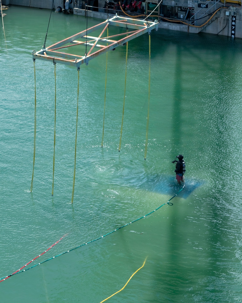 An aerial view of a waterway or basin shows a diver standing on or near a submerged object in green-tinted water. A large rectangular metal lifting frame is suspended above the water by yellow straps attached to a crane, with additional yellow rigging lines hanging down toward the water. The scene appears to be a salvage, recovery, or underwater inspection operation conducted from an industrial dock or quay.