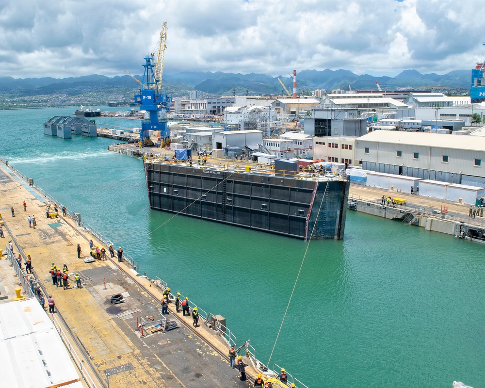 The image shows a naval shipyard facility, likely Pearl Harbor Naval Shipyard in Hawaii, based on the distinctive mountain backdrop and tropical setting. A large floating dry dock is being maneuvered into position between two concrete piers, with numerous workers in high-visibility safety gear observing and managing the operation. Industrial cranes, warehouses, and shipyard infrastructure are visible throughout the facility.