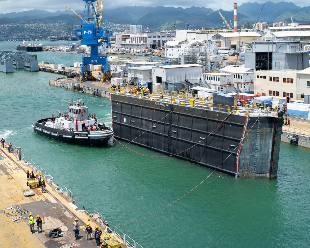 A large floating dry dock gate or caisson is being maneuvered by a tugboat in what appears to be a naval shipyard, likely Pearl Harbor Naval Shipyard in Hawaii based on the mountainous backdrop and tropical vegetation. Workers in high-visibility safety vests and hard hats are visible on the dock structure, the pier, and surrounding areas. The operation involves multiple personnel and mooring lines to position the massive steel structure.