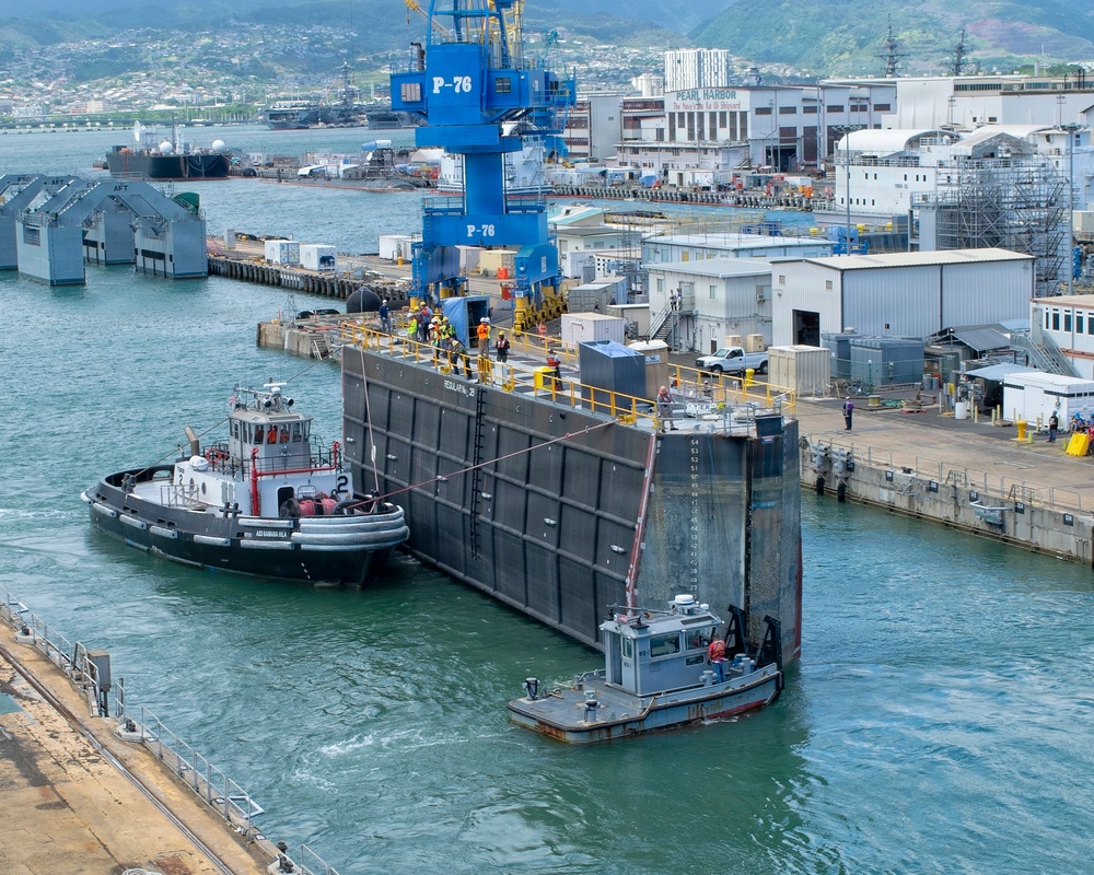 The image shows an active naval shipyard facility, identifiable as Pearl Harbor Naval Shipyard based on visible signage. A large floating dry dock caisson (gate) is being maneuvered by tugboats in the harbor channel, with workers in high-visibility safety gear visible on top of the structure. The blue crane labeled 'P-76' dominates the background, surrounded by industrial buildings, piers, and additional naval vessels.