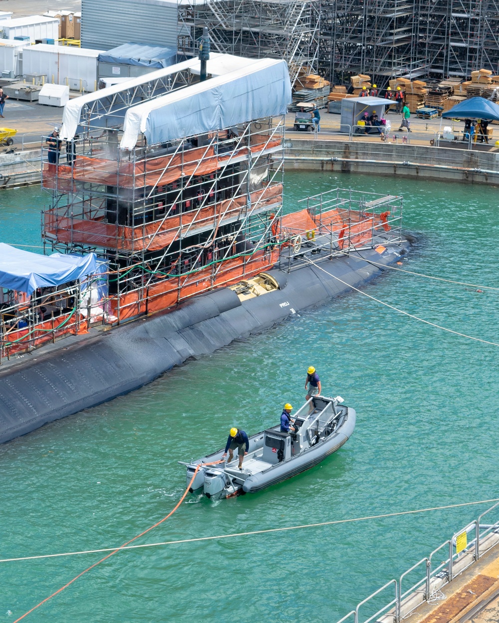 A submarine is docked at a naval shipyard undergoing maintenance or refit operations, with extensive scaffolding and orange safety netting surrounding its sail (conning tower). Workers in hard hats are visible both on the scaffolding and on a rigid inflatable boat (RIB) in the foreground, which appears to be handling mooring lines. The facility shows active industrial operations with storage areas, temporary structures, and multiple personnel on the pier.