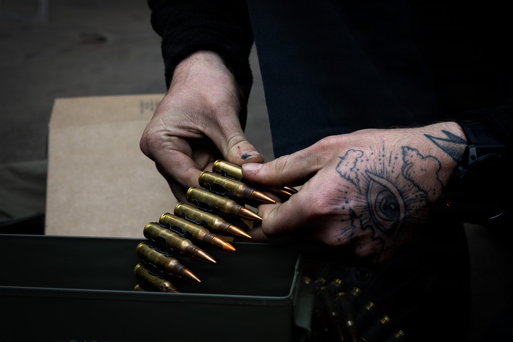 A person with a hand tattoo is handling a linked belt of rifle ammunition over a military-style metal ammo can. The rounds appear to be large-caliber brass-cased cartridges connected by a metallic disintegrating belt link system. A cardboard box is visible in the background.