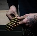 A person with a hand tattoo is handling a linked belt of rifle ammunition over a military-style metal ammo can. The rounds appear to be large-caliber brass-cased cartridges connected by a metallic disintegrating belt link system. A cardboard box is visible in the background.