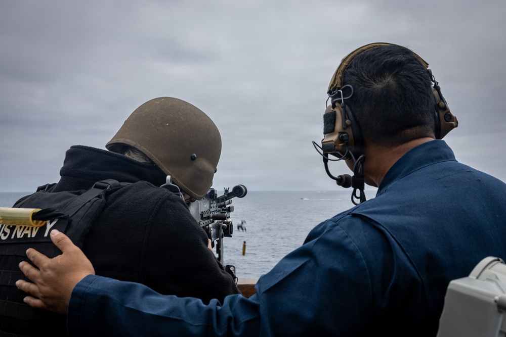 Two U.S. Navy personnel are positioned on the deck of a naval vessel at sea, with one sailor aiming a mounted machine gun toward the water. A second sailor wearing tactical hearing protection appears to be supervising or coaching the shooter. The overcast sky and open ocean suggest an at-sea training or live-fire exercise.