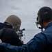 Two U.S. Navy personnel are positioned on the deck of a naval vessel at sea, with one sailor aiming a mounted machine gun toward the water. A second sailor wearing tactical hearing protection appears to be supervising or coaching the shooter. The overcast sky and open ocean suggest an at-sea training or live-fire exercise.