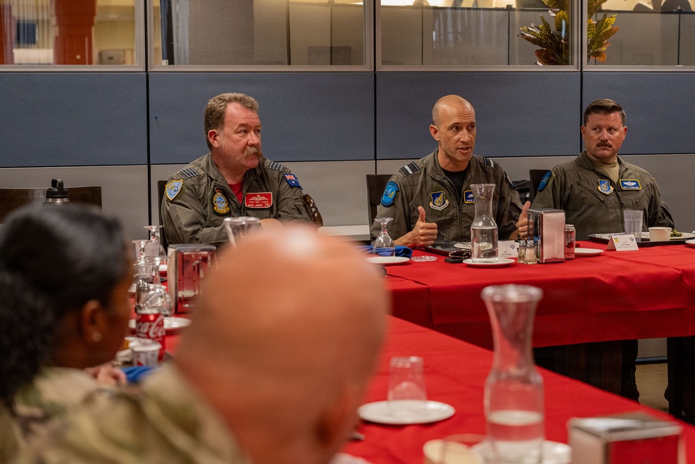 Three military personnel in flight suits are seated at a table with a red tablecloth, appearing to be engaged in a briefing or discussion during a meal event. The central figure is speaking and gesturing with his hands, while the others listen. The setting appears to be a military dining facility or conference room.