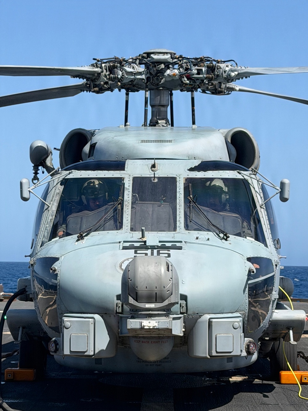 A front-facing view of a U.S. Navy military helicopter, numbered '516', parked on a ship's flight deck over open ocean. Two crew members in flight helmets are visible through the cockpit windscreen. The aircraft appears to be an MH-60 Seahawk variant equipped with a forward-looking sensor turret.