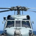 A front-facing view of a U.S. Navy military helicopter, numbered '516', parked on a ship's flight deck over open ocean. Two crew members in flight helmets are visible through the cockpit windscreen. The aircraft appears to be an MH-60 Seahawk variant equipped with a forward-looking sensor turret.