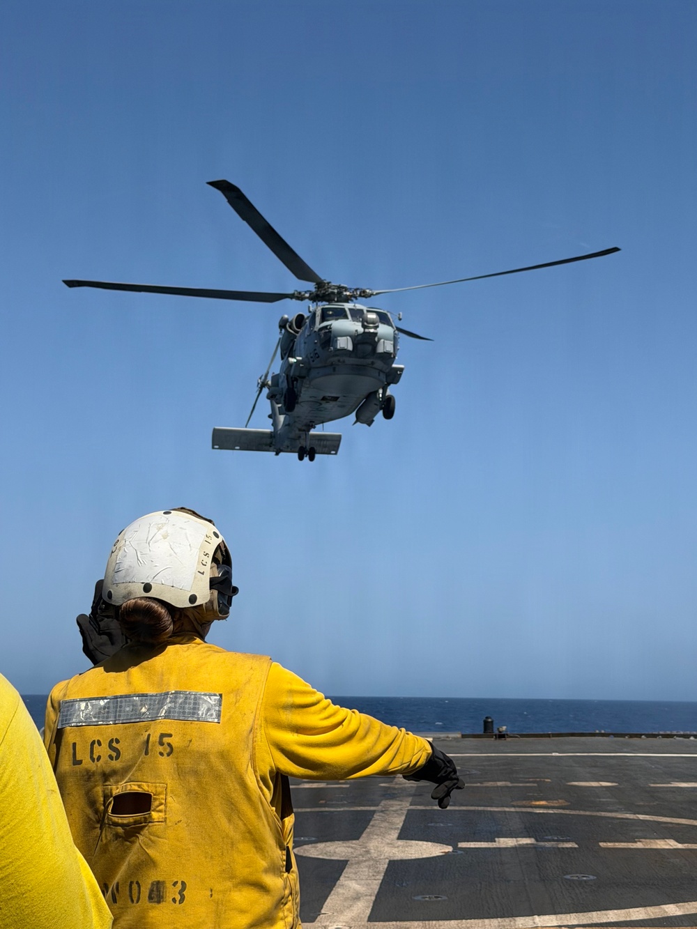 A U.S. Navy flight deck crew member aboard USS LCS 15 signals a military helicopter during a flight operations evolution on an open ocean flight deck. The crew member, wearing a yellow jersey and white helmet marked 'LCS 15', is directing what appears to be an MH-60 Seahawk helicopter hovering above the ship's flight deck. The scene takes place under clear blue skies over open ocean.