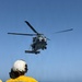 A U.S. Navy flight deck crew member aboard USS LCS 15 signals a military helicopter during a flight operations evolution on an open ocean flight deck. The crew member, wearing a yellow jersey and white helmet marked 'LCS 15', is directing what appears to be an MH-60 Seahawk helicopter hovering above the ship's flight deck. The scene takes place under clear blue skies over open ocean.