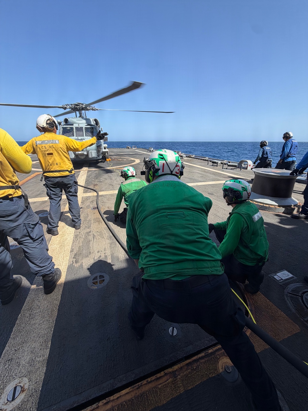 Naval personnel are conducting helicopter deck operations aboard what appears to be USS Billings (LCS-15), a U.S. Navy Littoral Combat Ship. Crew members in green jerseys and helmets are managing fuel hoses while a yellow-jersed flight deck coordinator signals to a military helicopter (likely an MH-60 Seahawk) on the flight deck. The operation is taking place at sea under clear blue skies.