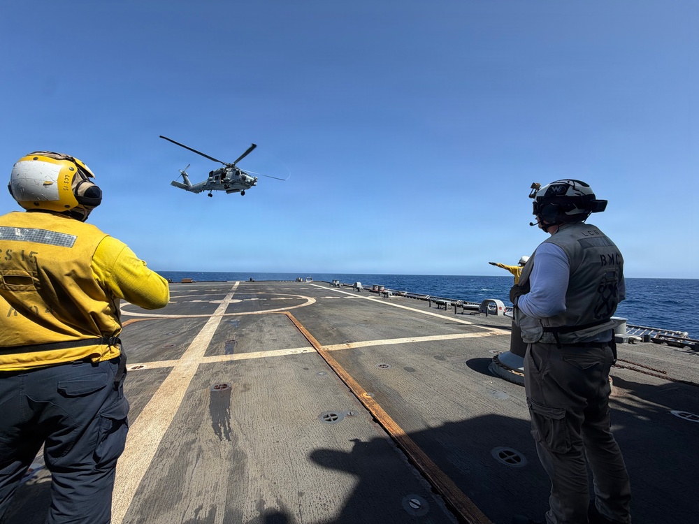 Two naval personnel are directing a military helicopter approaching or departing a ship's flight deck over open ocean. The personnel are wearing standard naval flight deck gear including helmets with hearing protection and high-visibility vests. The scene depicts a routine helicopter deck operations evolution aboard what appears to be a U.S. Navy vessel.