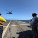 Two naval personnel are directing a military helicopter approaching or departing a ship's flight deck over open ocean. The personnel are wearing standard naval flight deck gear including helmets with hearing protection and high-visibility vests. The scene depicts a routine helicopter deck operations evolution aboard what appears to be a U.S. Navy vessel.